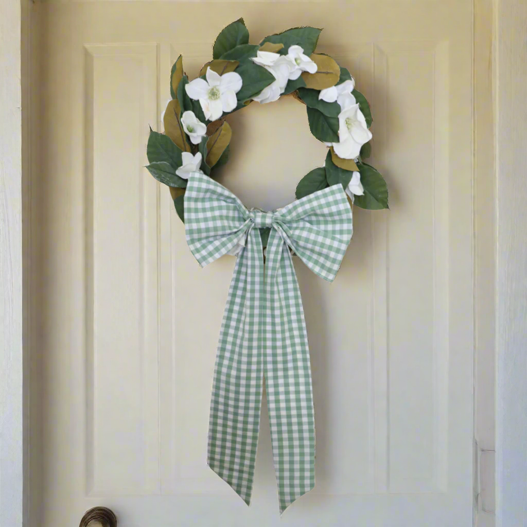Decorative wreath with green leaves and white flowers, tied with a green and white checkered ribbon on a white background.