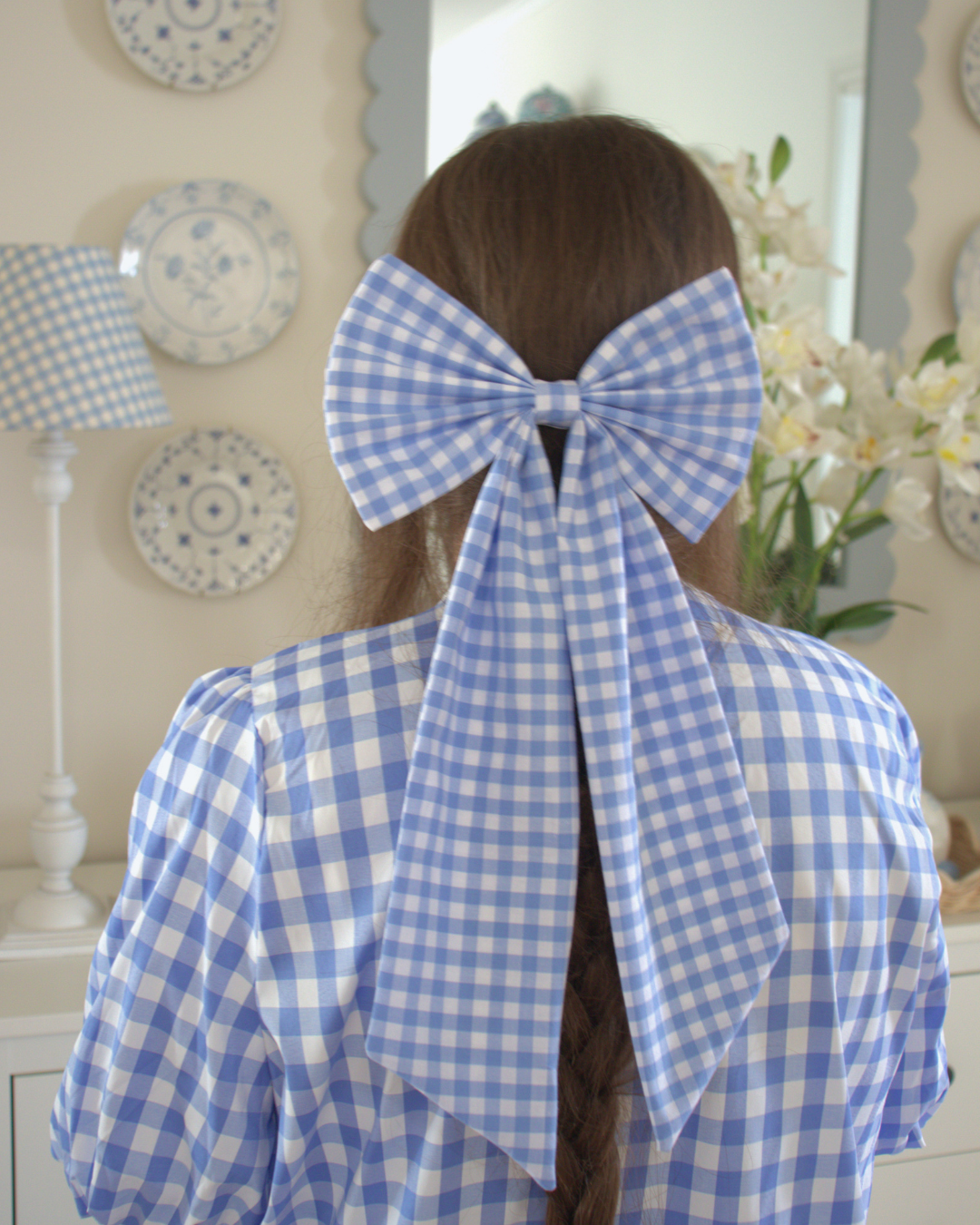 Person wearing a large blue and white checkered bow in a room with decorative plates and flowers.