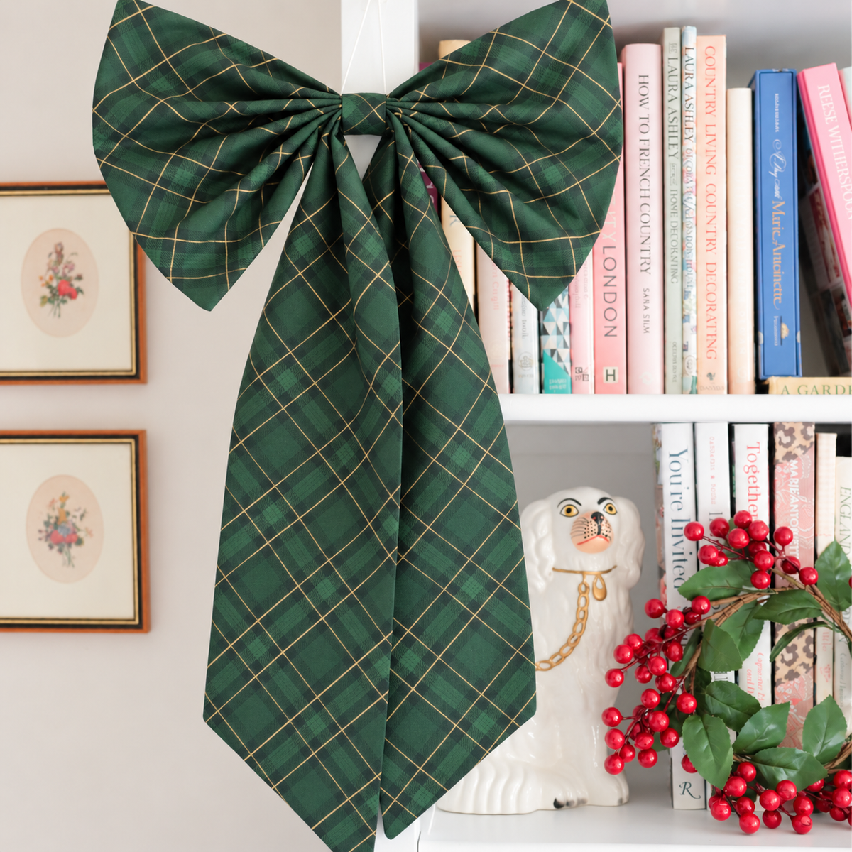 A large dark green bow with a gold plaid pattern tied around a white bookshelf, styled with framed art, books, a white dog statue, and red berries.