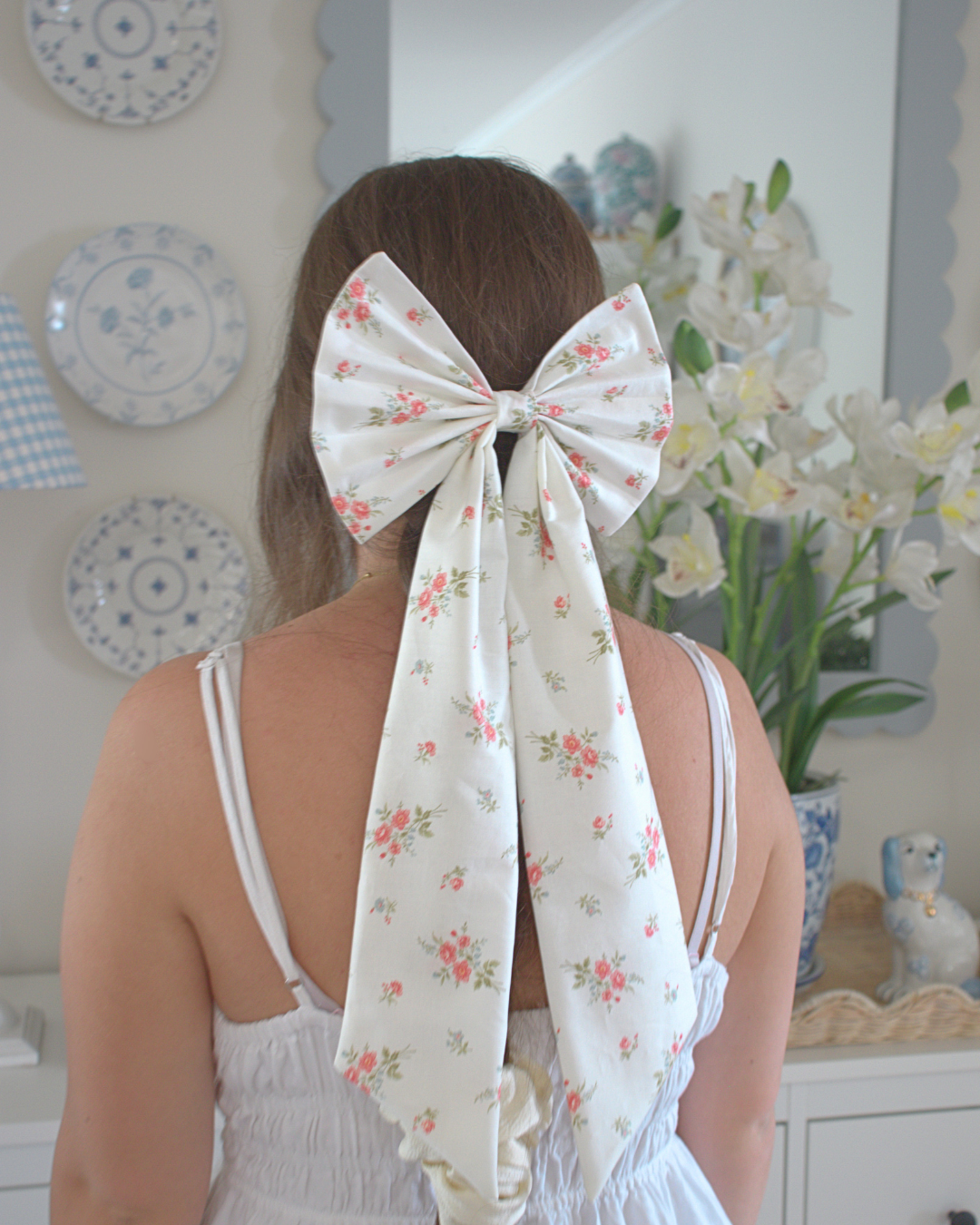 Person wearing a large floral bow in their hair with decorative plates and flowers in the background