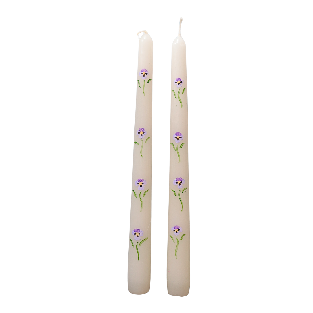 Two cream-colored taper candles decorated with small purple and white flowers and green stems, shown against a plain white background.