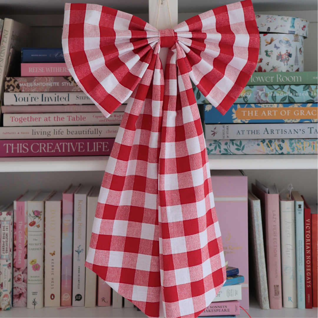 A large red and white gingham checkered bow made from fabric, hanging on a wooden bookshelf in front of books.