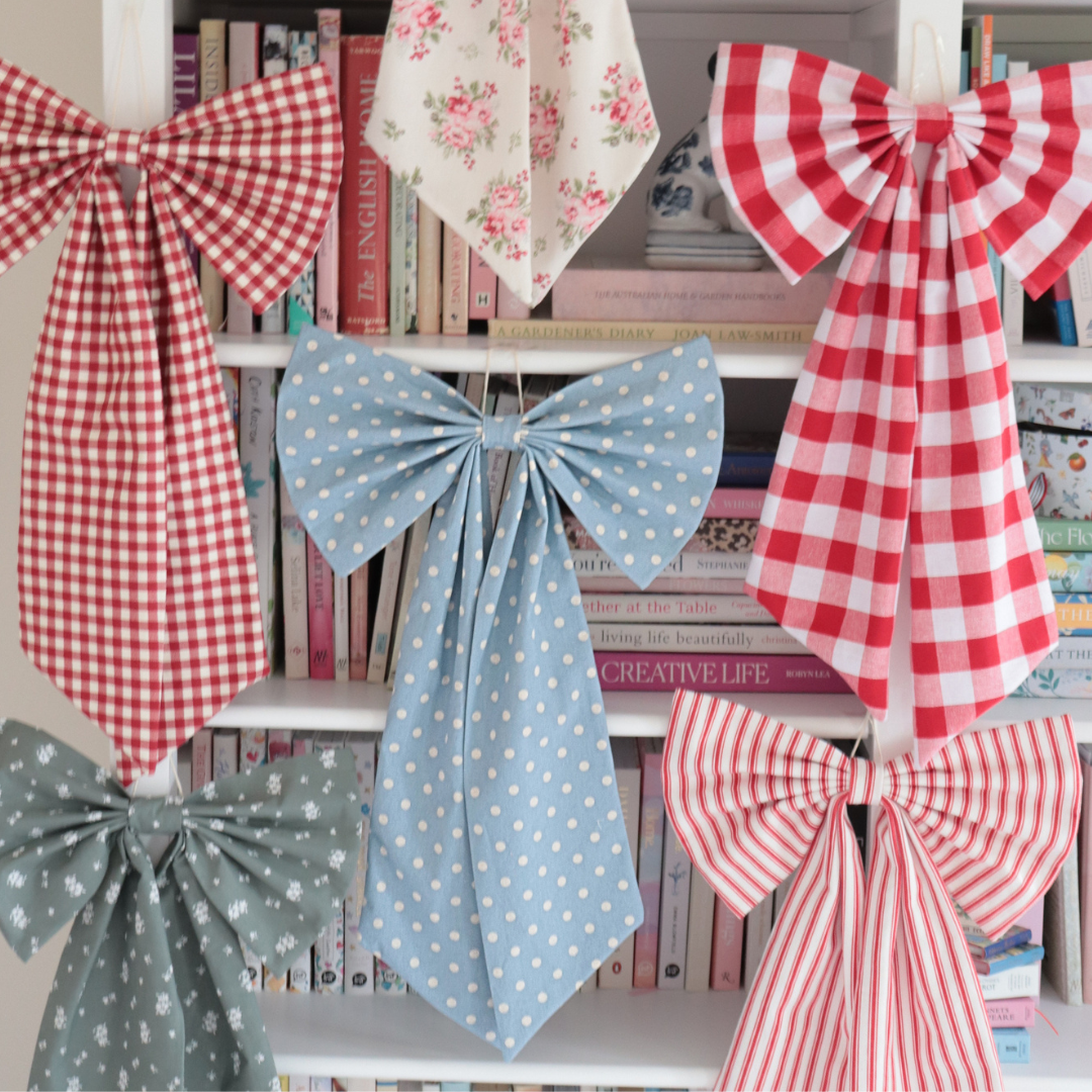 Six decorative fabric bows in various patterns (red gingham, light blue polka dots, red and white stripes, and a floral print) hanging on a white bookshelf.