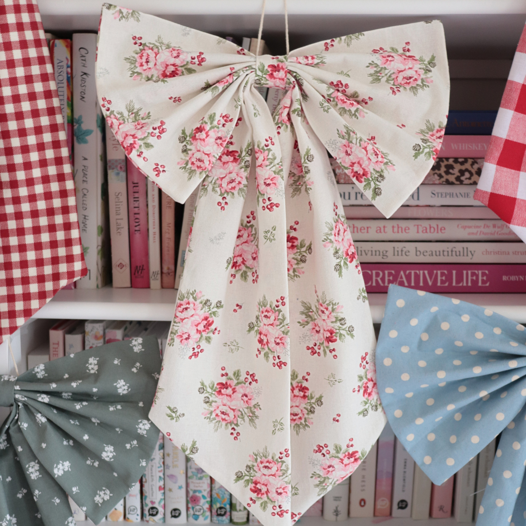 A large fabric bow with a pink and green floral print on an off-white background, hanging in front of a bookshelf filled with books and other decorative fabrics.
